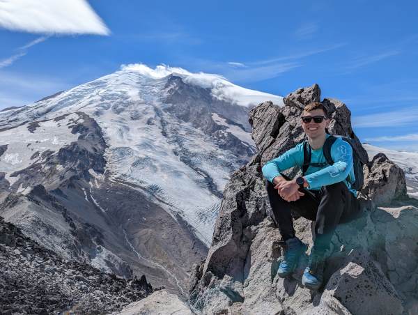 Photograph of Colton sitting on a rock structure with Mount Rainier in the background.